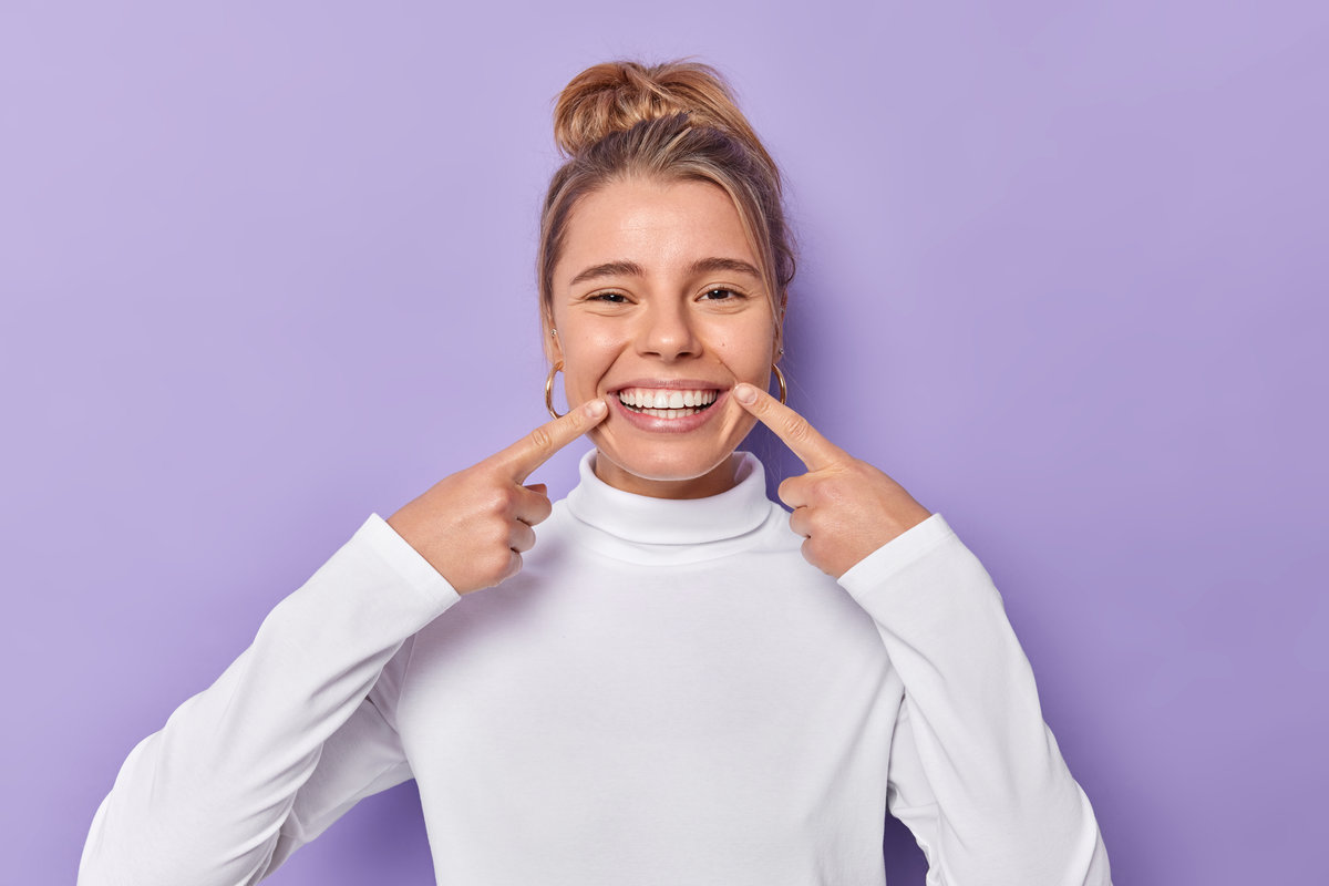 Dental care concept. Happy young fair haired woman points index fingers at toothy smile shows wel cared white teeth after whitening or dentist visit dressed casually isolated over purple background